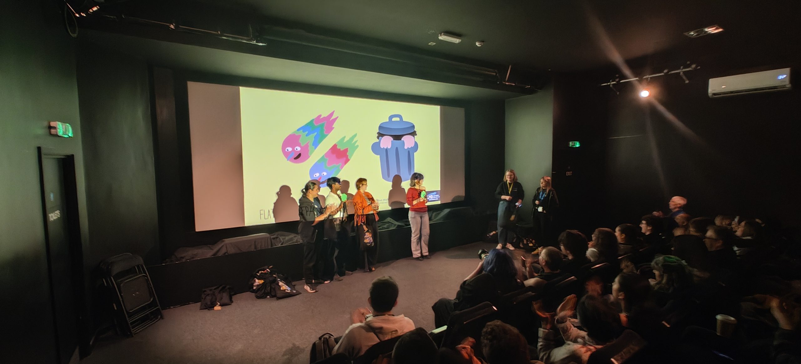 Young filmmakers standing at the front of a cinema to introduce their animated film, with a seated audience watching.