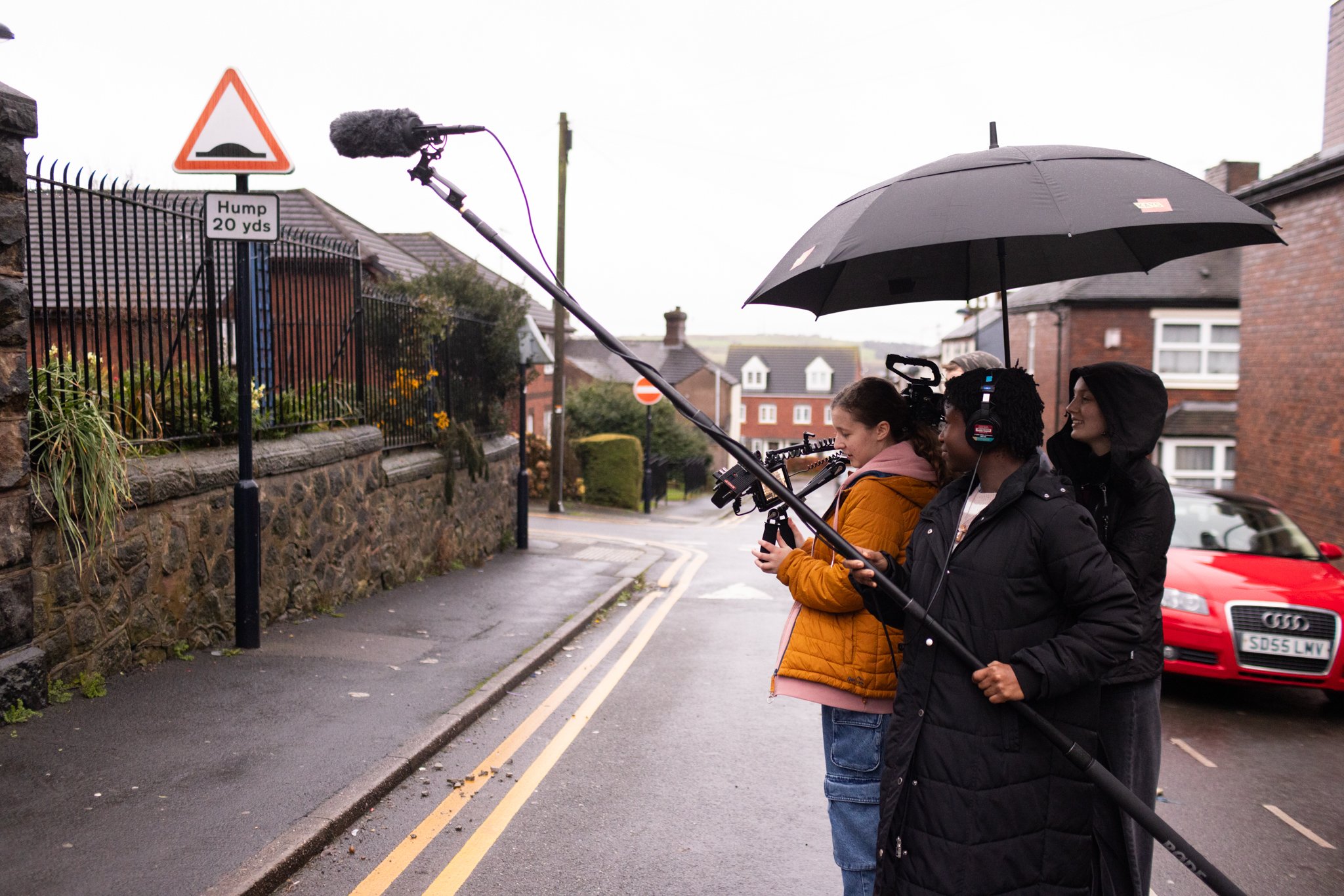 Three young BFI Film Academy crew members filming on a West Midlands street, one on camera, one on the boom, one holding an umbrella over the kit.