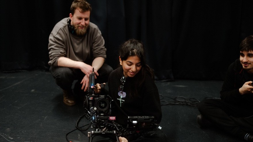 A young camera operator smiling behind a camera rig on a studio floor, with a tutor crouched beside her and another participant looking on.