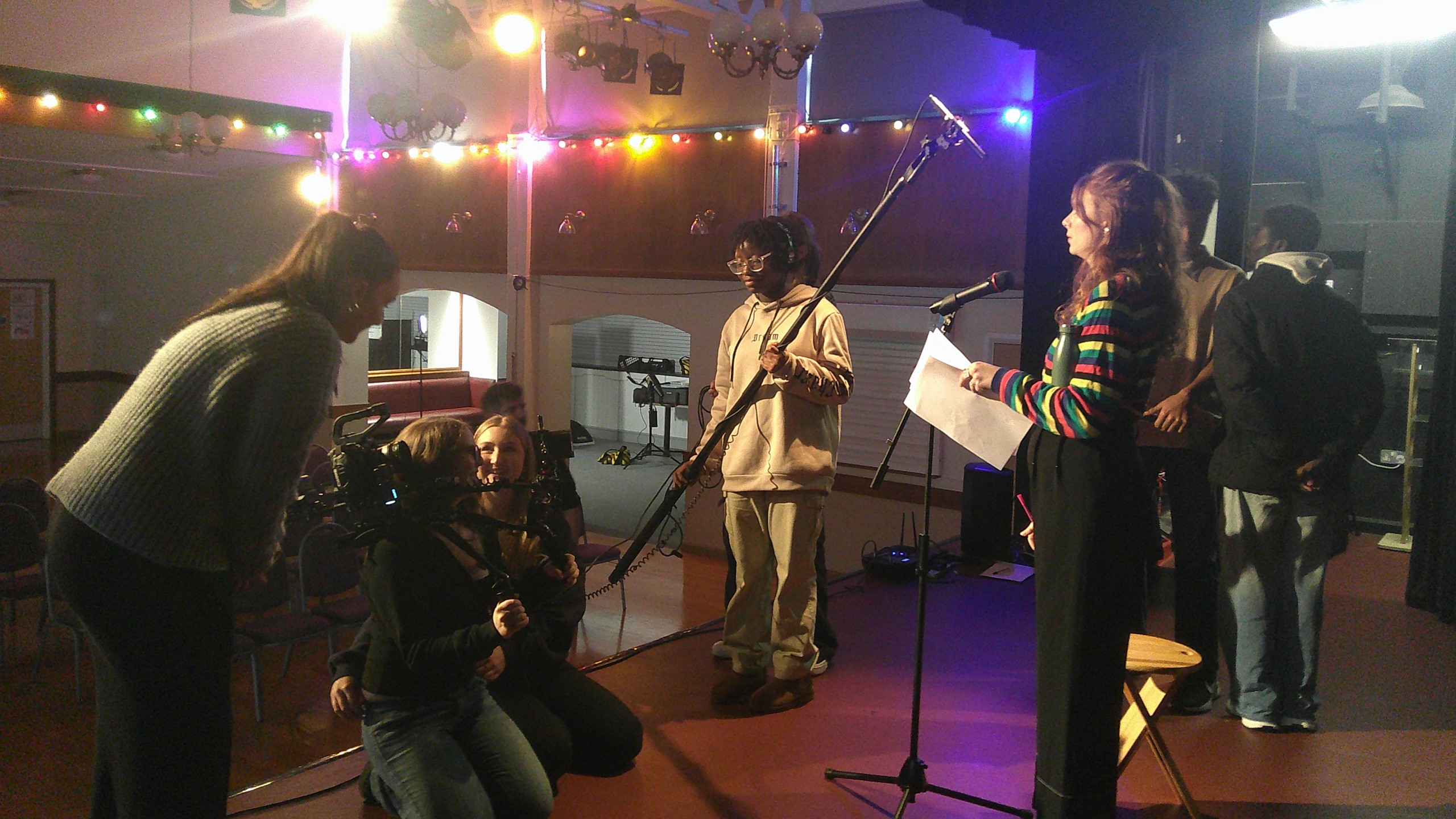 A BFI Film Academy crew at work in a lit hall: one kneeling behind a camera rig, one holding a boom microphone, and a presenter reading from a script at a mic stand.