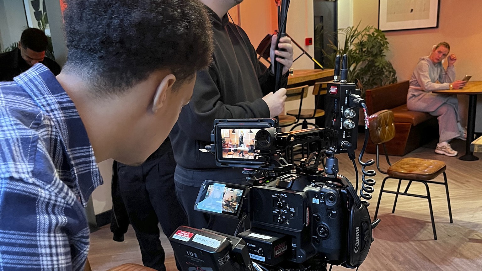 A young camera operator working a Canon cinema camera on a tripod in a cafe, with a crew member holding a boom microphone behind.